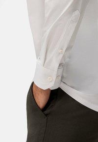 Witte blouse met lange mouwen en een knoopdetail bij de manchetten, gecombineerd met donkergrijze broek. Close-up toont de textuur van de stof en het ontwerp van de manchetten.