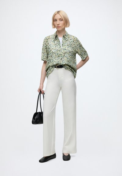 Woman with short blonde hair wearing a green floral short-sleeve shirt, white wide-leg pants, black shoes, holding a black handbag.