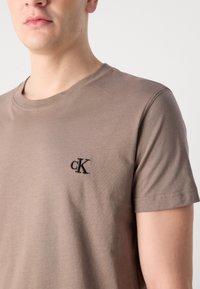Smeđa pamučna majica s okruglim izrezom, kratkim rukavima i izvezenim "cK" logom na lijevoj strani prsnog koša. Glatka tekstura i prilagođeni dizajn.