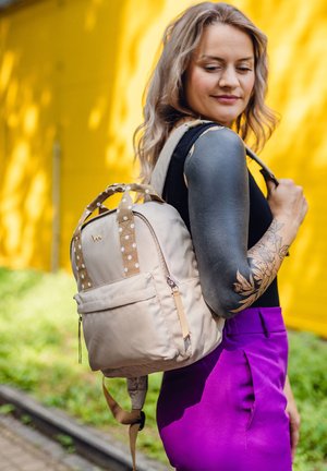 Beige Canvas-Rucksack mit gepunkteten Trägern, Reißverschluss-Fronttasche und matter Oberfläche. Das Design ist kompakt und strukturiert.