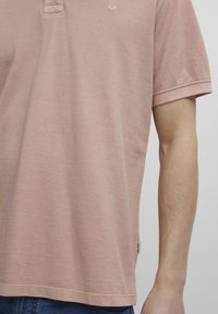 Lyserosa polo-skjorte laget av teksturert bomull. Har en liten krage, korte ermer og et merke på kanten.