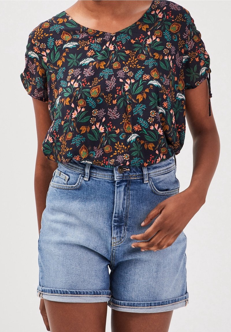 Mujer lleva una blusa corta de manga con estampado floral oscuro, metida en unos pantalones cortos de mezclilla azul claro con el dobladillo vuelto, con una mano descansando cerca del bolsillo.