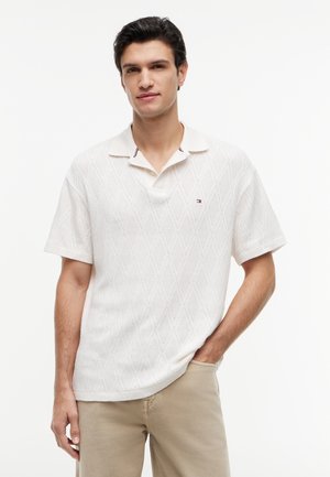 Junger Mann mit dunklen Haaren, der ein weißes, kurzärmeliges, strukturiertes Poloshirt und beige Hosen trägt und mit einer Hand in der Hosentasche steht.