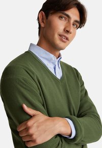 Zielony sweter w serek z miękkiej dzianiny, za łożony na jasnoniebieską koszulę z guzikami i kołnierzykiem. Ręce skrzyżowane w zrelaksowanej pozycji.