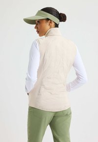 Beige kviltad väst med hög krage, kombinerad med en långärmad vit topp och olivgröna byxor, samt en matchande grön skärmmössa.