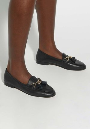 Black leather loafers with a flat sole, featuring a gold hardware accent and tassels on the front. Smooth texture with rounded toe.