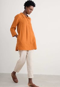 Losse oranje linnen tuniek met een kraag en knopen, gecombineerd met lichtbeige linnen broek en bruine klompen.
