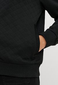 Čierny prešívaný sweatshirt s textúrovaným povrchom, rebrovanými manžetami a bočnou kapsou. Materiál vyzerá mäkko a teplo.