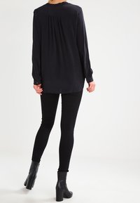 Svart blus med långa ärmar och veckad baksida, samt knäppningar vid handleden, tillsammans med svarta skinnyjeans och ankelboots, på en neutral bakgrund.