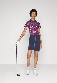 Golfový outfit pozostáva z vzorovanej ružovo-námorníckej polo košele, námorníckej sukne s bielymi akcentmi a bielych golfových topánok. Golfová palica je držaná v ľavej ruke.
