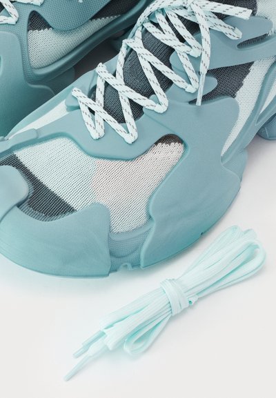 Turquoise athletic shoes with mesh and rubber overlays, featuring textured, curved designs and matching laces beside them.