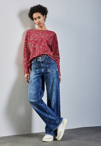 Pull à motifs rouges avec une coupe ample, associé à un jean bleu à jambes larges. Le modèle porte des baskets blanches, se tenant contre un mur gris clair.