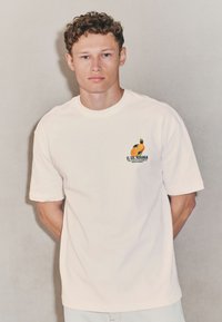 T-shirt beige chiaro in cotone con maniche corte, scollo rotondo e grafica frontale di arance con la scritta "EL SOL NARANJA."