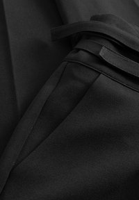 Közeli kép fekete anyagból készült nadrágról, oldalzsebbel és hozzáillő, fekete szövetből készült övvel, amely egy hurokba van kötve.