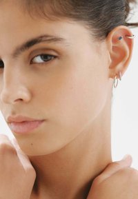 Aros de plata en forma de aro y un pendiente azul adornan la oreja, mostrando un diseño minimalista sobre un tono de piel neutro. Textura suave, estilo elegante.