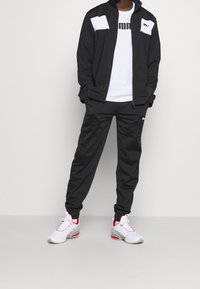 Chándal ligero negro con acentos blancos, que incluye una chaqueta con cremallera y pantalones ajustados, combinado con zapatillas deportivas blancas con detalles rojos.
