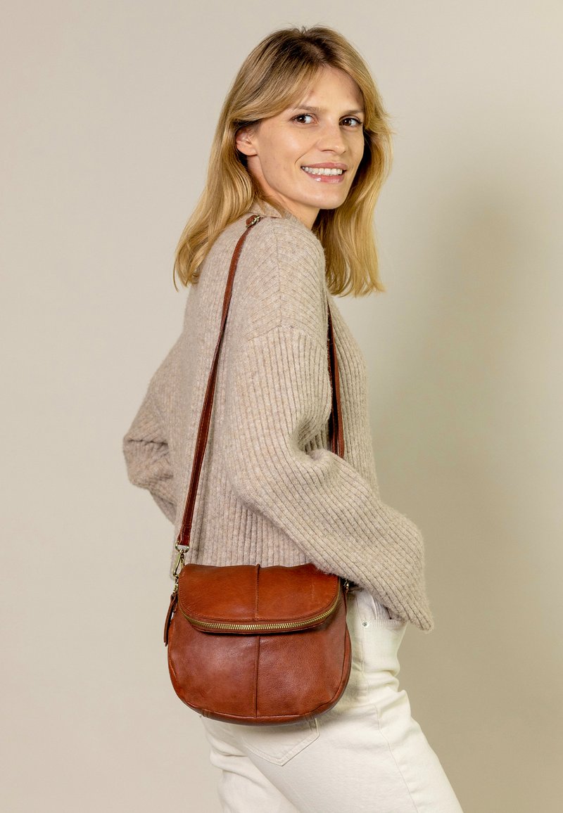 Hnědá kožená crossbody taška se zipem, s kulatým tvarem a jemnými švy. Nosit s pleteným béžovým svetrem.