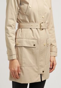 Trench-coat beige ceinturé avec boutons dorés et grandes poches avant, porté sur un pantalon noir, visible de la mi-cuisse aux épaules.