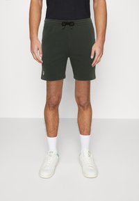 Donkergroene katoenen shorts met een verstelbare touwkoord taille en logo patch. G gedragen met witte sneakers en sokken. Eenvoudig ontwerp, tot halverwege de dijen.