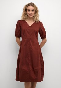 Rudų spalvų, iki kelių siekiantis suknelė su V formos iškirpte, pūstomis rankovėmis ir juosmens įtemptu dizainu. Pagaminta iš lygios medžiagos, turinčios subtilų blizgesį.