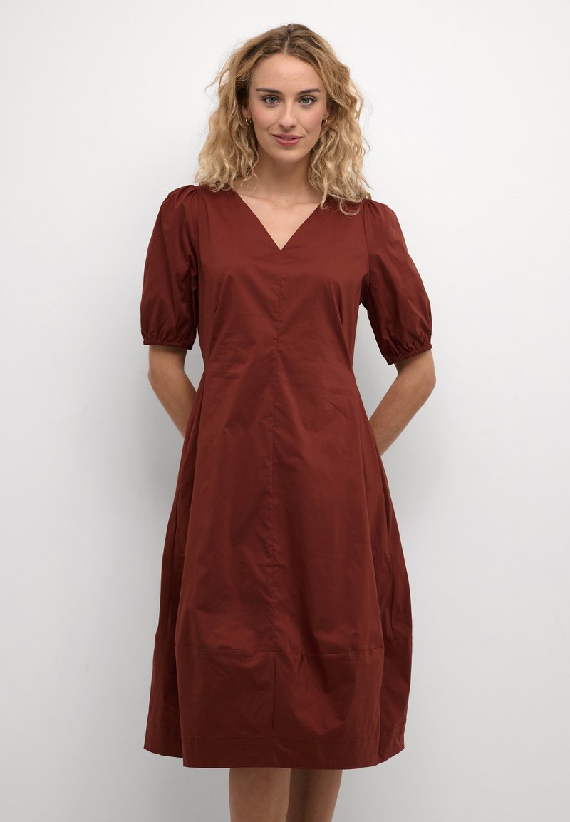 Rudų spalvų, iki kelių siekiantis suknelė su V formos iškirpte, pūstomis rankovėmis ir juosmens įtemptu dizainu. Pagaminta iš lygios medžiagos, turinčios subtilų blizgesį.