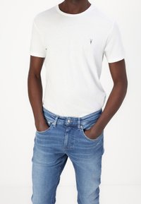 Vit kortärmad T-shirt med rund halsringning, med en liten logotyp på bröstet. Buren med ljusblå jeans, händerna i fickorna.