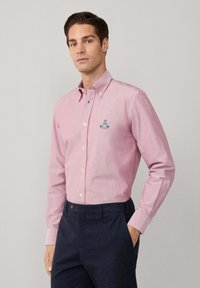Hombre vestido con una camisa de botones de manga larga rosa con un pequeño logo en el pecho, metida en pantalones navy oscuros, de pie con una mano en el bolsillo.