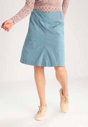 Femme portant une jupe bleu clair mi-longue, un haut à manches longues en dentelle rose pâle et des baskets beige, debout sur un fond blanc.