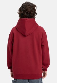 Red hoodie with a large, adjustable hood, ribbed cuffs, and hem; soft fabric with a smooth texture, designed for a relaxed fit.