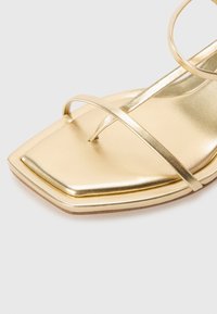Sandalo metallico dorato con punta quadrata, sottili cinturini, texture liscia e suola piatta con un delicato bordo decorativo.