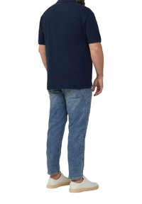 Mężczyzna stojący tyłem, ubrany w granatową koszulkę polo, jasnoniebieskie dżinsy i białe trampki na białym tle.