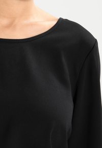 Haut noir à manches longues avec un large décolleté en forme de coupe et une texture lisse, avec des coutures visibles le long des bords.