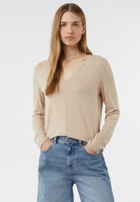 Beige langärmliges V-Ausschnitt-Oberteil mit einer weichen Textur, entspannter Passform und Manschetten. Kombiniert mit hellblauen hochgeschnittenen Jeans.