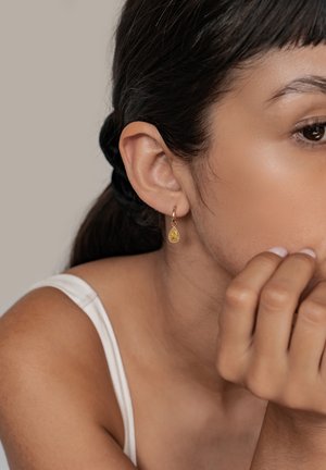 Goldene Creolen mit einem birnenförmigen, gelben Stein, die um den Stein herum mit filigranen Details verziert sind und an einem Ohr eines Models getragen werden.