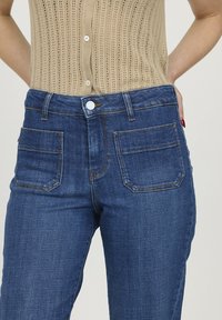 Jean en denim bleu foncé avec taille haute, dotés de cinq poches et de coutures contrastantes. Le tissu semble lisse avec un minimum d'usure.