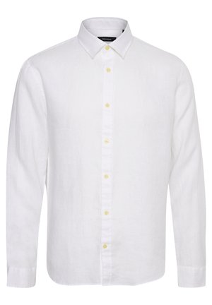 Homme portant une chemise blanche à manches longues boutonnée, un pantalon noir et des sandales marron à double bride, debout devant un fond uni.
