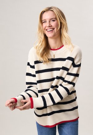 Lachende vrouw met lang blond haar, gekleed in een crèmekleurige trui met zwarte horizontale strepen en rode rand, die haar mouw verstel.