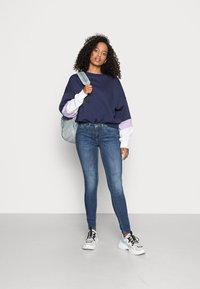 Sweatshirt oversized azul marinho com mangas brancas e lilás claro, combinado com jeans ajustados azuis e ténis brancos chunky, segurando uma mochila cinza.