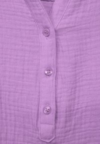 Camisa de botones lavanda con una tela texturada y arrugada. Presenta un escote en forma de V y cuatro botones redondos en la parte frontal.