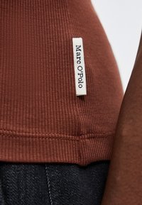 Gros plan de l'ourlet d'une chemise en tissu côtelé marron avec une étiquette blanche de la marque Marc O'Polo cousue sur le côté, associée à un jean en denim foncé.