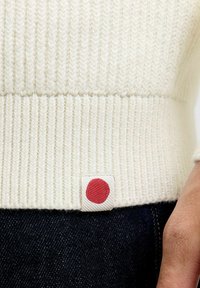 Maglione a maglia di colore crema, con un ampio orlo a costine e un'etichetta rossa nell'angolo in basso. La texture appare morbida e calda.