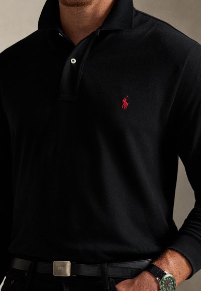 Muž nosiaci čierne polo s dlhým rukávom s červeným vyšívaným logom na hrudi, čierny opasok a náramkové hodinky so zeleným ciferníkom.