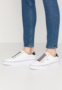 Baskets blanches décontractées avec des œillets dorés, des accents marine et un panneau en tissu rayé. Présentent une semelle texturée et des détails cousus.