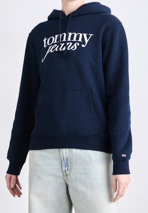 Osoba ubrana w granatową bluzę z kapturem z białym napisem „tommy jeans” oraz jasnoniebieskie dżinsy, ukazana od szyi do bioder.