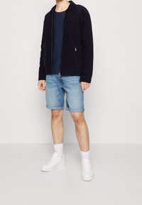 Navy corduroy takki kauluksella, yhdistetty kevyisiin sinisiin farkkushortseihin ja valkoisiin lenkkareihin. Mallilla on navy T-paita alla.