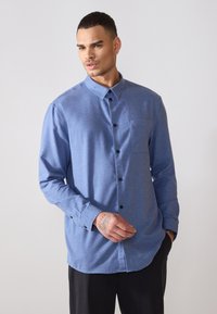 Chemise bleu à manches longues, boutonnée, en tissu texturé, avec des boutons noirs et une poche poitrine, ayant une coupe décontractée et un ourlet arrondi.