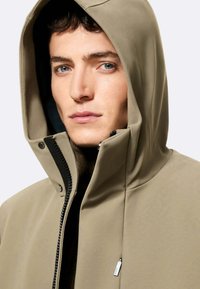 Beige gewatteerde jas met capuchon, ritssluiting, zacht materiaal en contrasterende zwarte voering. Voorzien van een klein zijzakje met ritssluiting en een minimalistisch ontwerp.