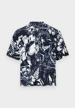 Ung person med krøllet hår iført en kortærmet skjorte med blomsterprint, hvid tanktop, grå shorts, stripede sokker og sorte ankelstøvler.