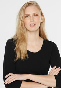Schwarzes Langarmshirt mit Rundhalsausschnitt, glatter Textur und taillierter Passform. Modell mit glattem, schulterlangem blonden Haar.
