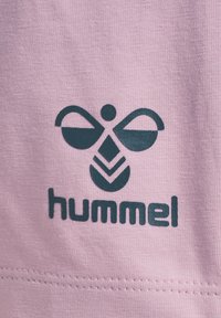 Lichtroze stof met een donkergroen Hummel-logo met een gestileerd bijontwerp en tekst. Glad oppervlak, korte stiksels zichtbaar.